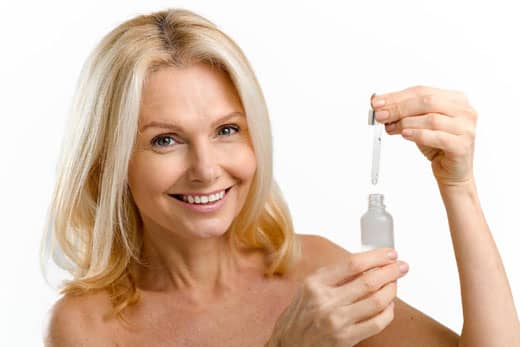 Sophisticated middle aged woman applies regenerating serum on the face. Charming blonde 40s lady using anti-aging complex with hyaluronic acid isolated on white background